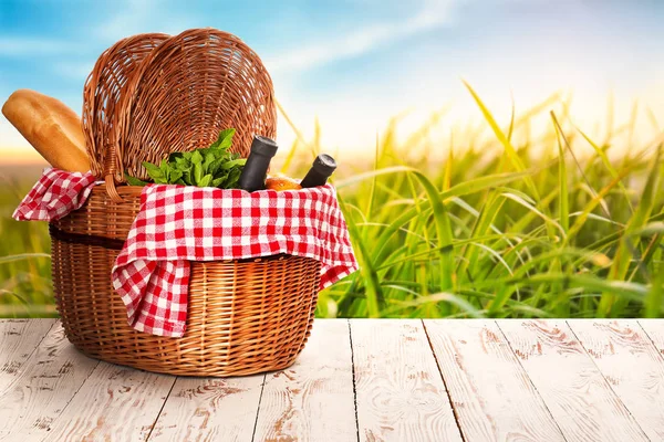 Плетеная корзина с вкусной едой и напитками для пикника на деревянном столе возле пышной зеленой травы — стоковое фото