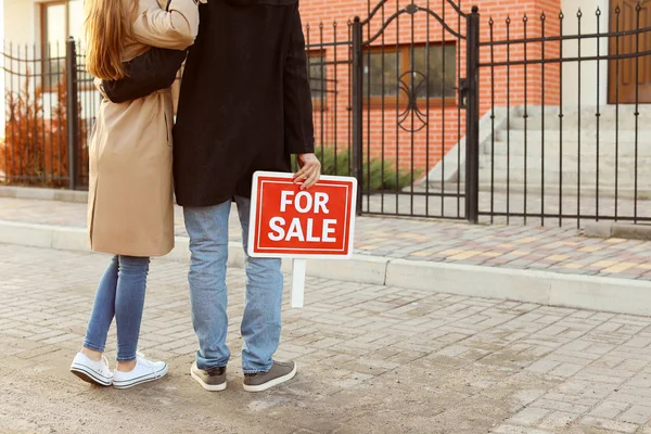 Casal jovem com placa de venda perto de sua casa — Fotografia de Stock