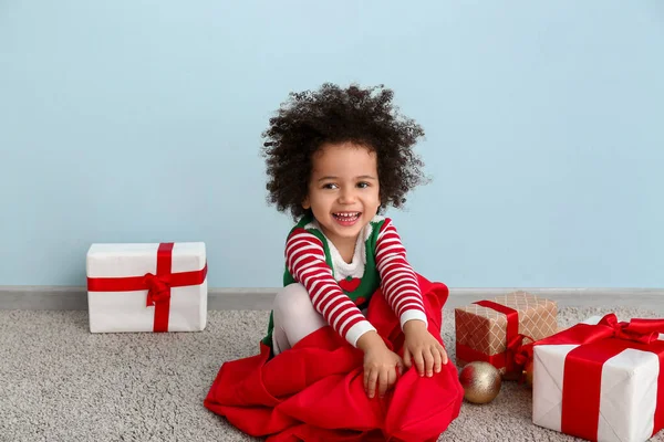 Menina em traje de elfo, com saco de Papai Noel e presentes de Natal sentados perto da parede de cor — Fotografia de Stock