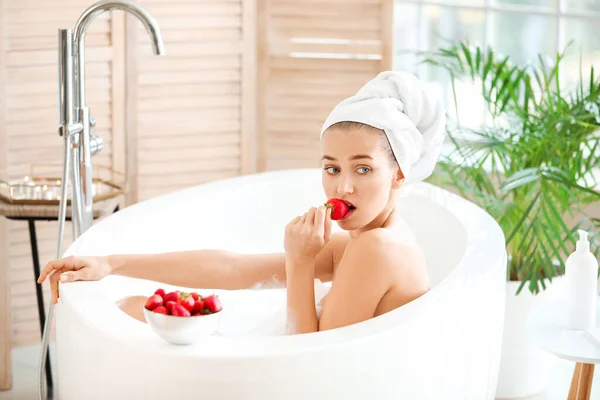 Beautiful young woman eating sweet strawberries in bathroom — Stock Photo, Image
