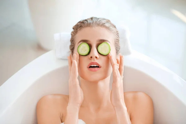 Hermosa mujer joven con rodajas de pepino tumbado en el baño —  Fotos de Stock