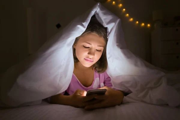 Tiener meisje met mobiele telefoon onder deken 's nachts — Stockfoto