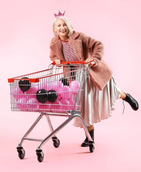 Крутая модная зрелая женщина с воздушными шарами в корзине для покупок на цветном фоне — стоковое фото