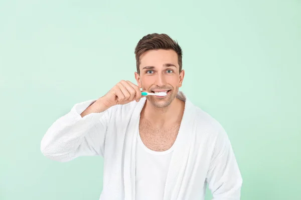 Handsome man in bathrobe cleaning teeth on color background