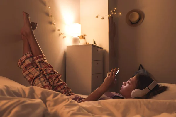Teini-ikäinen tyttö, jolla on matkapuhelin kuuntelemassa musiikkia, kun makaa sängyssä yöllä — kuvapankkivalokuva