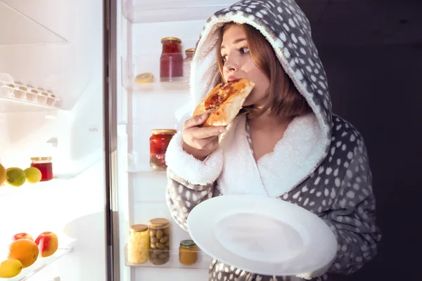 Девочка-подросток ест нездоровую пищу возле холодильника ночью — стоковое фото