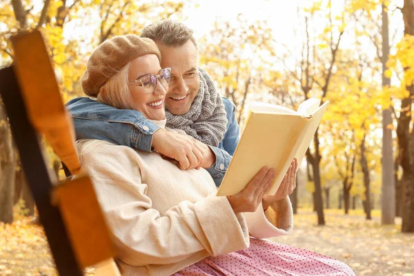 Счастливая взрослая пара читает книгу, сидя на скамейке в осеннем парке — стоковое фото