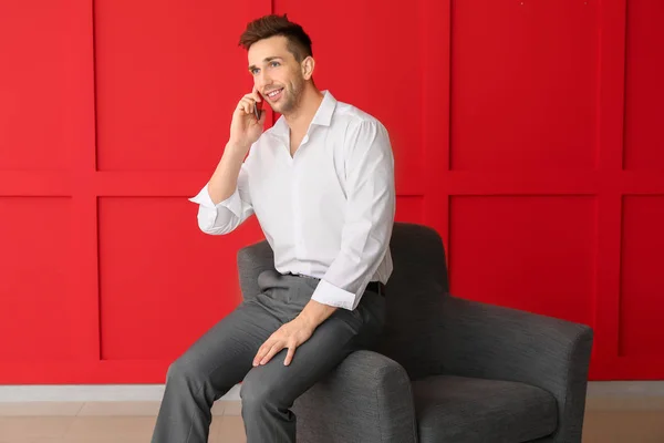 Hombre de negocios guapo hablando por teléfono móvil mientras está sentado en el sillón cerca de la pared de color —  Fotos de Stock