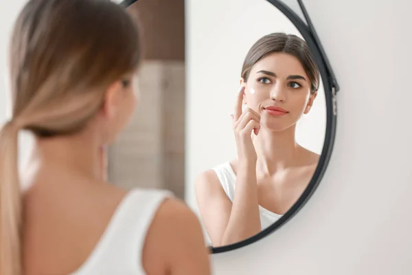 Hermosa mujer joven aplicando crema en su cara cerca del espejo en el baño — Foto de Stock