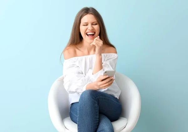 Happy woman with mobile phone sitting in armchair on color background