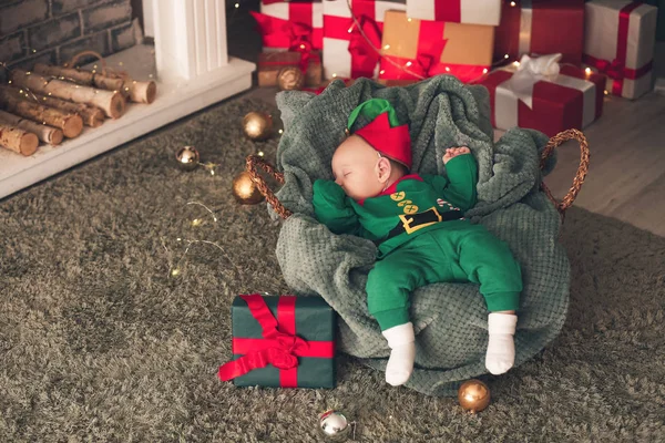 Pequeno bebê em traje de duende e com presente de Natal dormindo em cesta em casa — Fotografia de Stock