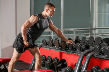 Spor salonunda dambıllarla spor yapan kaslı bir adam.