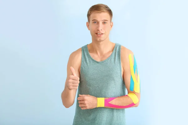 Uomo sportivo con nastro fisioterapico applicato sul braccio mostrando il gesto del pollice verso l'alto sullo sfondo chiaro — Foto Stock