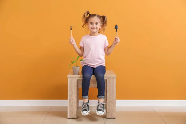 Cute little girl z młodą rośliną w doniczkach i narzędzi siedzi w pobliżu ściany kolor — Zdjęcie stockowe