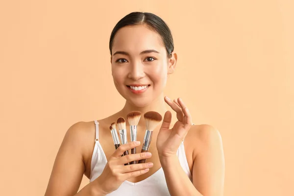 Portrait of young Asian woman with makeup brushes on color background — Stock Photo, Image