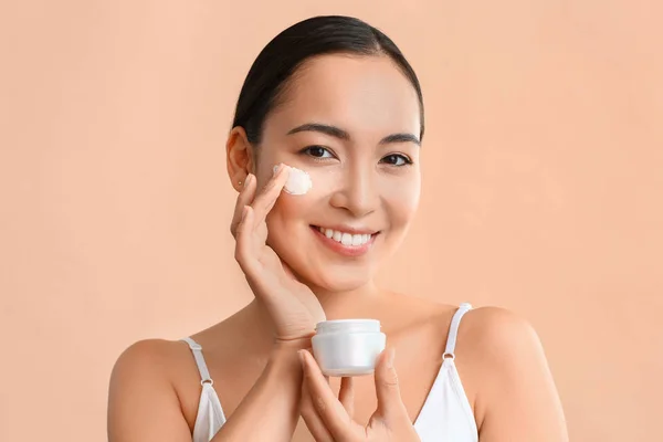 Beautiful Asian woman applying cream against color background — Stock Photo, Image