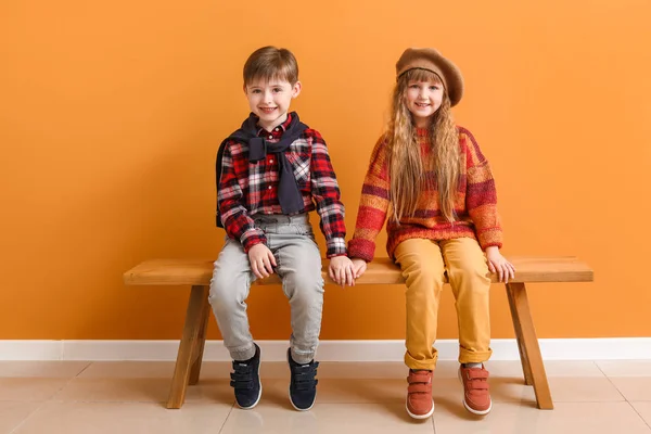 Cute little children in autumn clothes sitting on bench near color wall — Stock Photo, Image