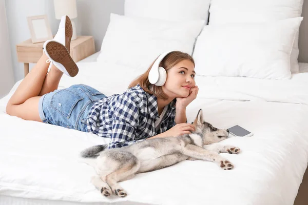 Cute teenage girl listening to music with funny husky puppy on bed at home — Stock Photo, Image