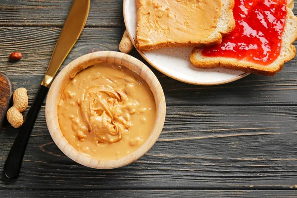 Manteiga de amendoim saborosa na tigela e pão fresco na mesa de madeira — Fotografia de Stock