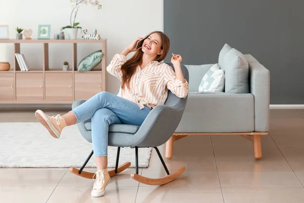 Beautiful woman talking by mobile phone while sitting in armchair at home — Stock Photo, Image