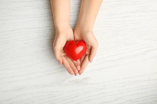 Mani femminili con cuore rosso su sfondo bianco in legno. Concetto di cardiologia — Foto Stock