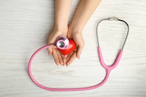 Female hands with red heart and stethoscope on white wooden background. Cardiology concept — Stock Photo, Image