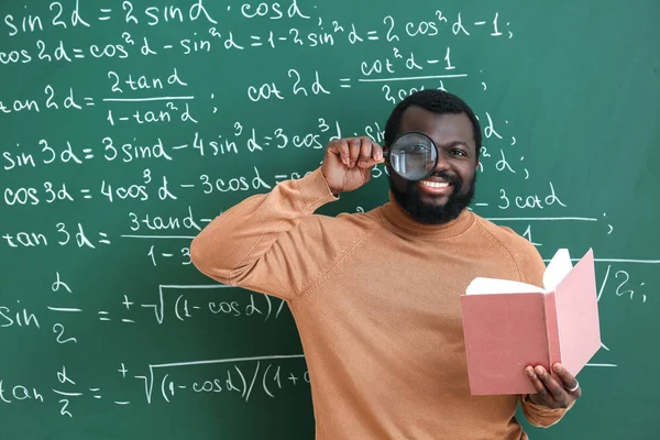 African-American math teacher with book and magnifier near blackboard in classroom