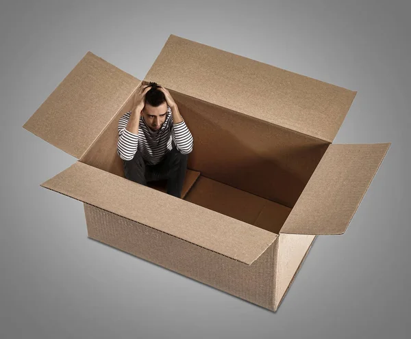 Depresivní mladý muž sedí v lepenkové krabici na šedém pozadí — Stock fotografie
