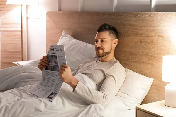 Bonito homem lendo jornal enquanto estava deitado na cama à noite — Fotografia de Stock