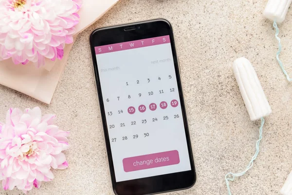 Menstrual calendar on screen of mobile phone and feminine products on grey background