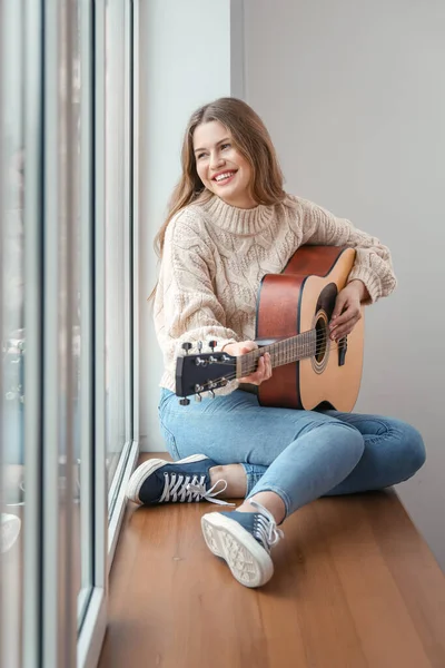 Young woman with guitar sitting on window sill at home — Stockfoto