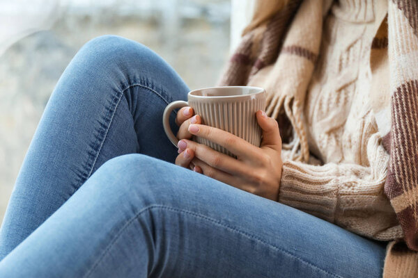 Young woman drinking hot tea on window sill at home, closeup