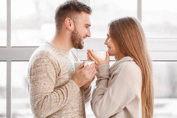 Pareja feliz con tazas de té caliente descansando cerca de la ventana en casa — Foto de Stock