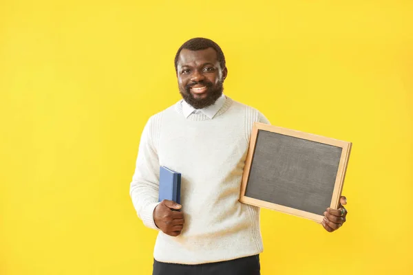 African-American teacher with chalkboard and book on color background — 스톡 사진