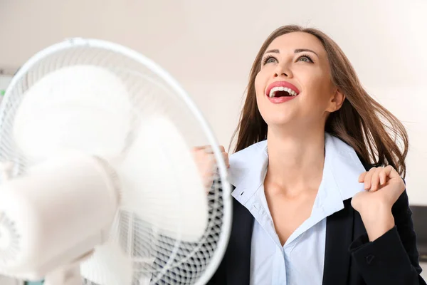 Young woman using electric fan during heatwave in office — Stock Photo, Image