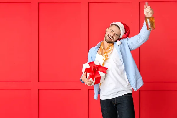 Pijany człowiek w kapeluszu Świętego Mikołaja, prezent i szampan na tle koloru — Zdjęcie stockowe