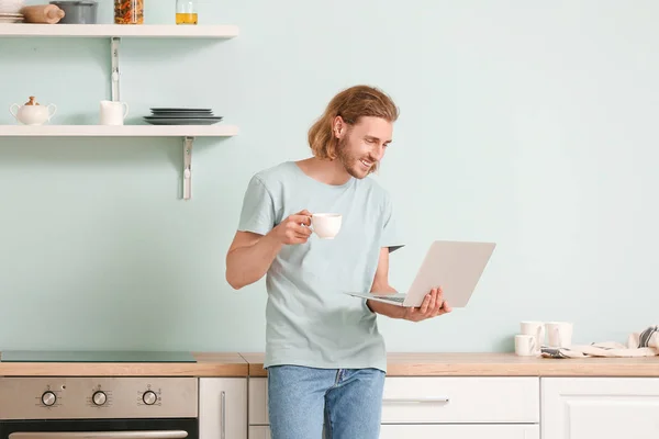 Handsome man with laptop drinking coffee in kitchen at home — Stock Photo, Image