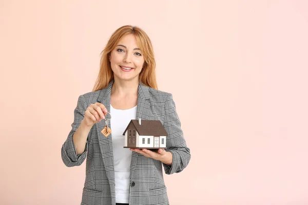 Female real estate agent with key and model of house on color background — Stock Photo, Image