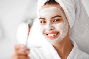 Güzel genç bir kadın banyoya yüz maskesi takıyor.