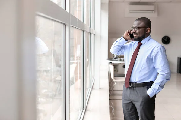 Hombre de negocios afroamericano guapo hablando por teléfono móvil cerca de la ventana en la oficina — Foto de Stock