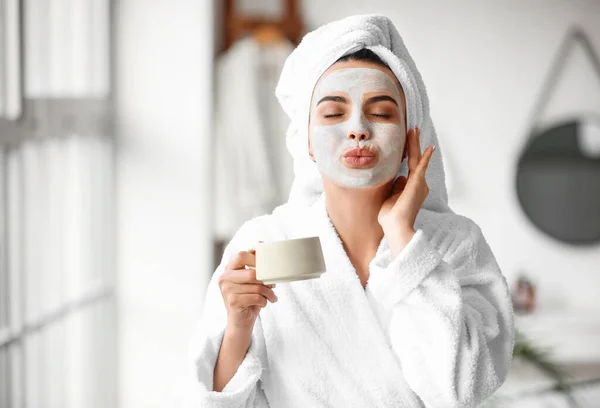 Schöne junge Frau mit Gesichtsmaske trinkt Kaffee im Badezimmer — Stockfoto