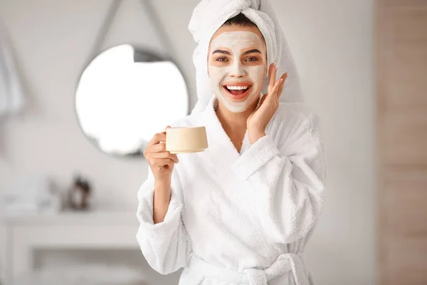 Beautiful young woman with facial mask drinking coffee in bathroom — Stock Photo, Image