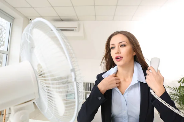 Young woman using electric fan during heatwave in office — Stock Photo, Image