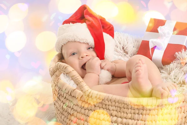 Cute little baby with Santa Claus hat and gift lying in basket against blurred Christmas lights — Stock Photo, Image