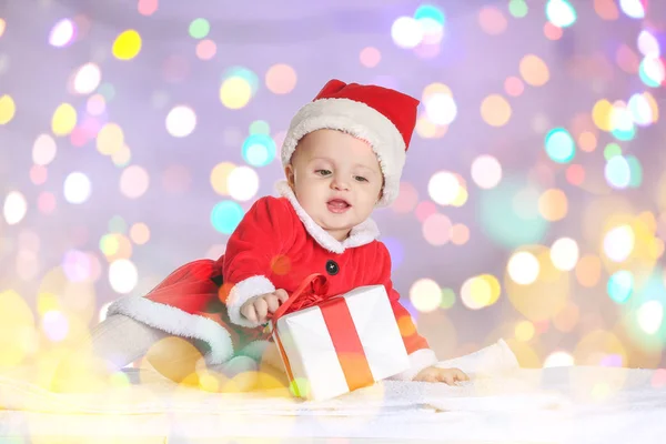 Cute little baby in Santa Claus costume and with Christmas gift against blurred lights — Stock Photo, Image