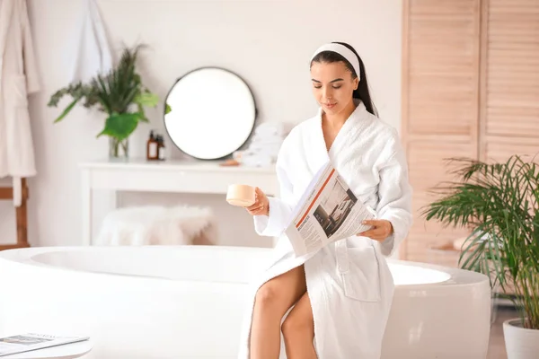 Beautiful young woman drinking coffee while reading newspaper in bathroom — Stock Photo, Image