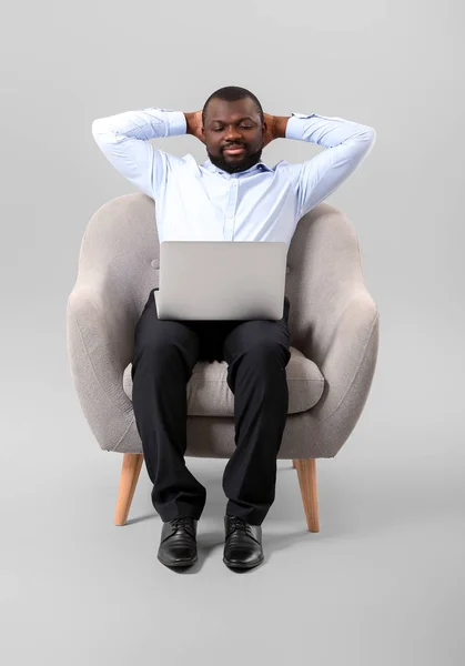 Афроамериканец с ноутбуком сидит в кресле на сером фоне — стоковое фото
