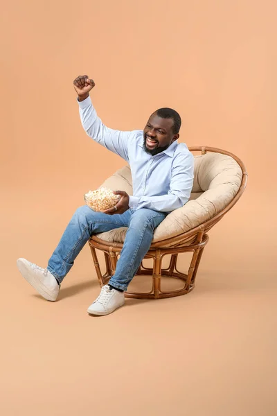 Emotionaler afrikanisch-amerikanischer Mann mit Popcorn vor dem Fernseher, während er im Sessel vor farbigem Hintergrund sitzt — Stockfoto