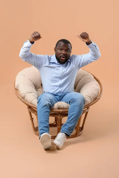 Emotional African-American man watching TV while sitting in armchair against color background — Stock Photo, Image