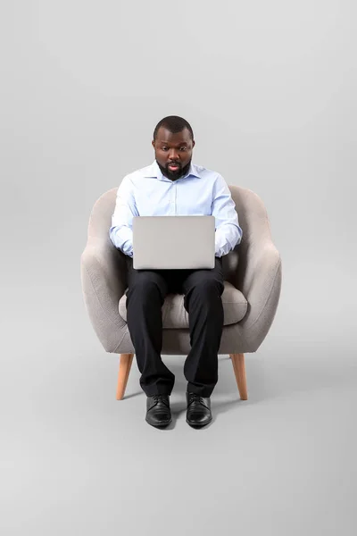 African-American man with laptop sitting in armchair against grey background — Stock Photo, Image